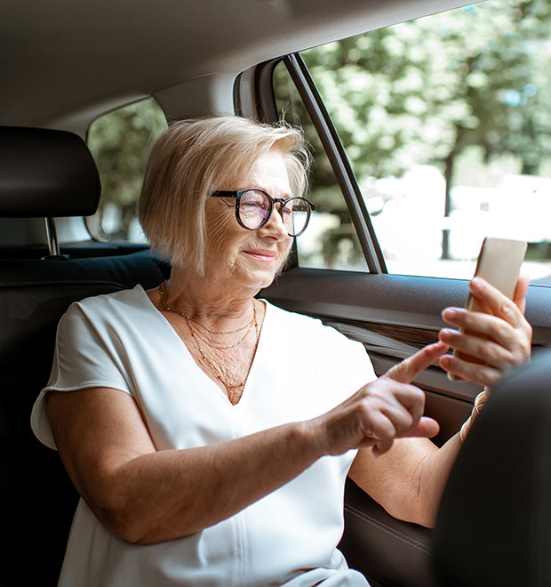 Older woman in back-row car seat checking her finances from her mobile phone. 
