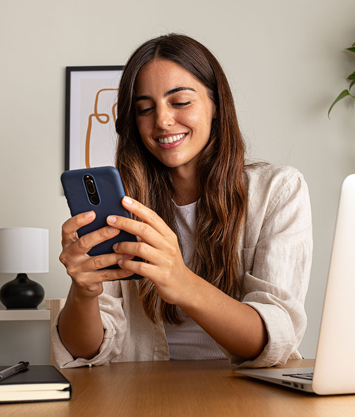 Image of a woman smiling while looking at a cell phone she&rsquo;s holding close to her face. She is at home with a laptop beside her.