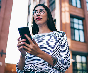 A woman outdoors, looking away from her phone with a thoughtful expression, as if deciding what to do.